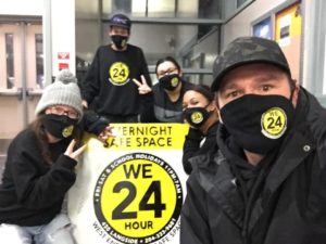 group of people with masks on standing around a sign that says WE24